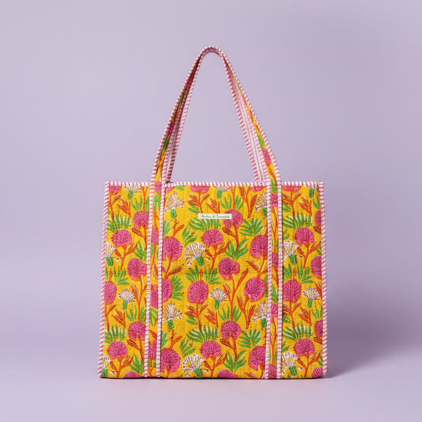 A colorful tote bag with a yellow background, pink and green floral pattern, and pink-and-white striped handles, displayed on a wooden stand inside a shop.