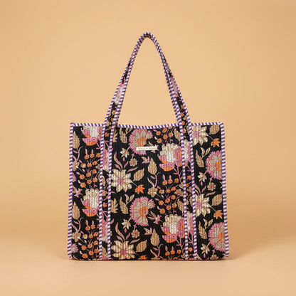 A large tote bag with a black background and colorful floral embroidery, hanging on a wooden display stand inside a toy and home goods store.