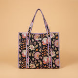 A large tote bag with a black background and colorful floral embroidery, hanging on a wooden display stand inside a toy and home goods store.