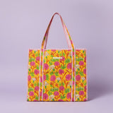 A colorful tote bag with a yellow background, pink and green floral pattern, and pink-and-white striped handles, displayed on a wooden stand inside a shop.