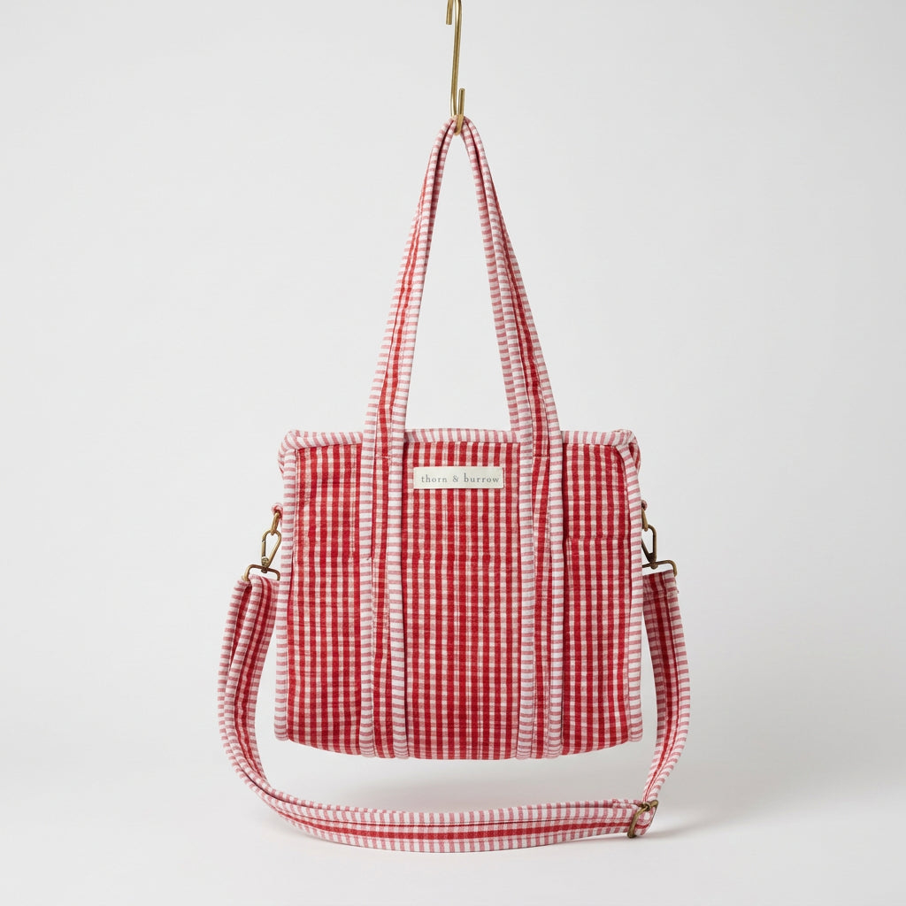 A red and white gingham tote bag with a matching shoulder strap, hanging from a yellow hook in a retail shop display.