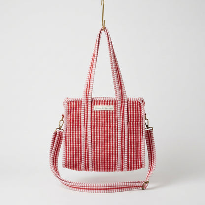 A red and white gingham tote bag with a matching shoulder strap, hanging from a yellow hook in a retail shop display.