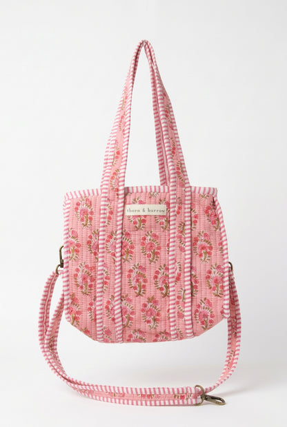 A pink floral tote bag with a striped shoulder strap, hanging from a white metal display stand inside a retail store.