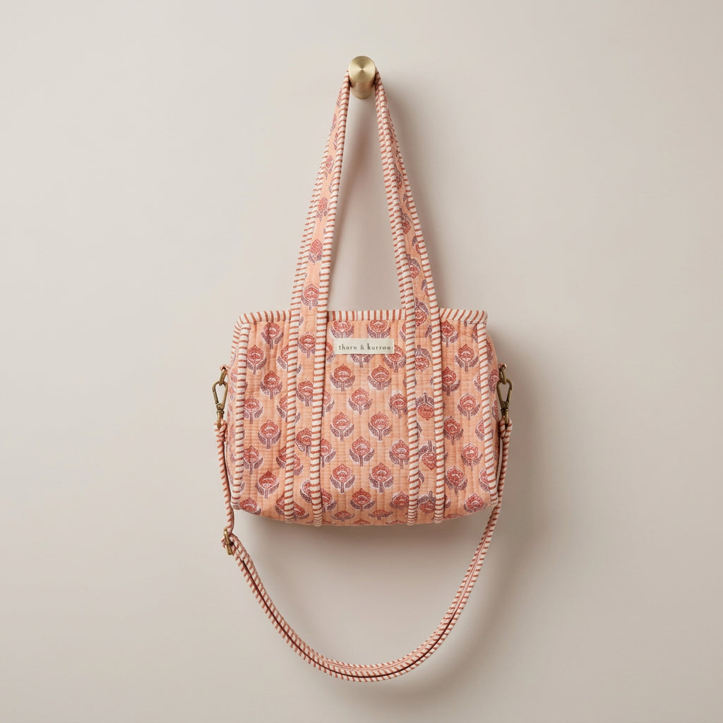 A patterned tote bag with long shoulder straps hangs from a yellow hook inside a white arched display frame in a retail store. The bag has a warm beige base with a repeating red, pink, and blue floral/geometric print, and a small white label on the front.