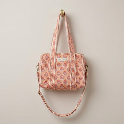 A patterned tote bag with long shoulder straps hangs from a yellow hook inside a white arched display frame in a retail store. The bag has a warm beige base with a repeating red, pink, and blue floral/geometric print, and a small white label on the front.