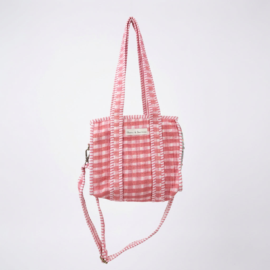 A pink and white gingham tote bag with a red heart-shaped pocket, hanging from a white metal arch in a retail store.