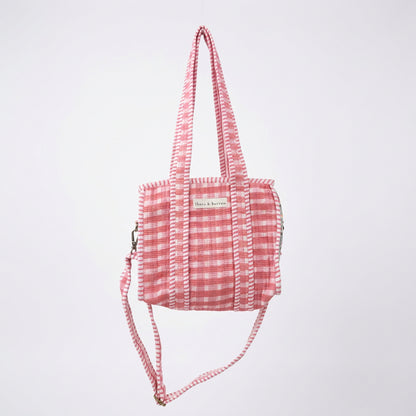 A pink and white gingham tote bag with a red heart-shaped pocket, hanging from a white metal arch in a retail store.