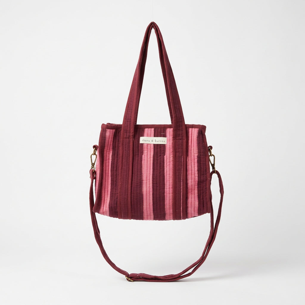 A burgundy velvet tote bag with vertical pink stripes and a matching shoulder strap, hanging from a white metal display frame inside a retail store.