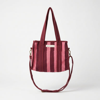 A burgundy velvet tote bag with vertical pink stripes and a matching shoulder strap, hanging from a white metal display frame inside a retail store.