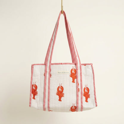 A woven tote bag with red and white striped handles and red lobster motifs, hanging in a retail store display.