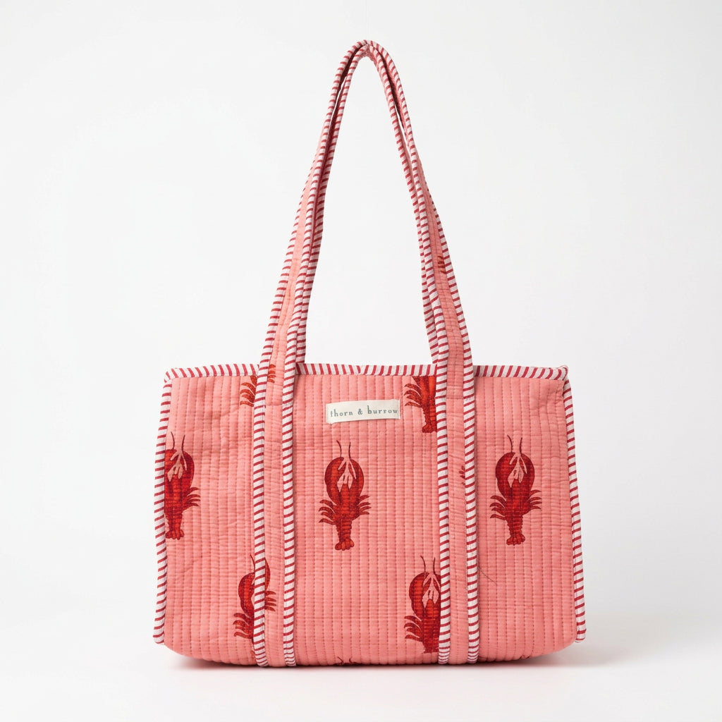 A pink woven tote bag with red lobster motifs and striped handles, hanging in a retail store setting.