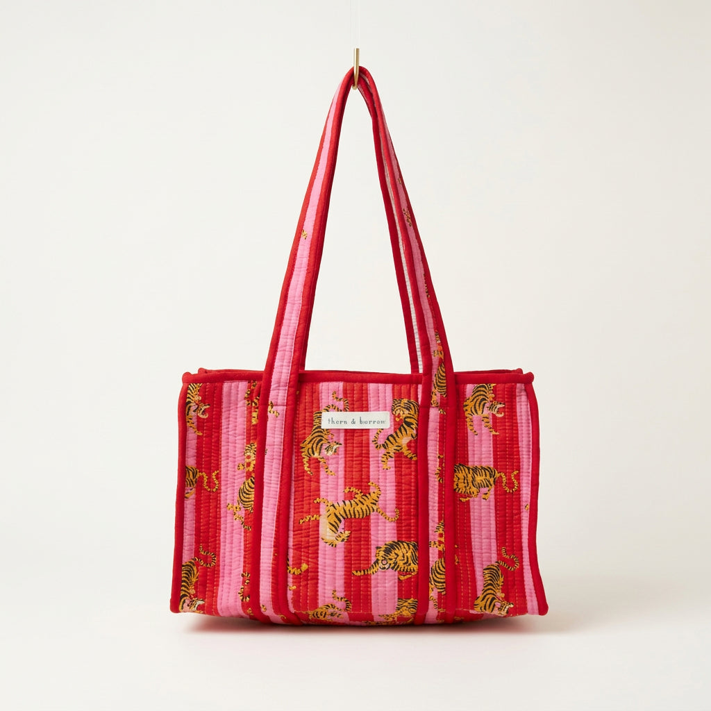 A red woven tote bag with a pink and yellow tiger print pattern, hanging from a gold hook in a retail store display.