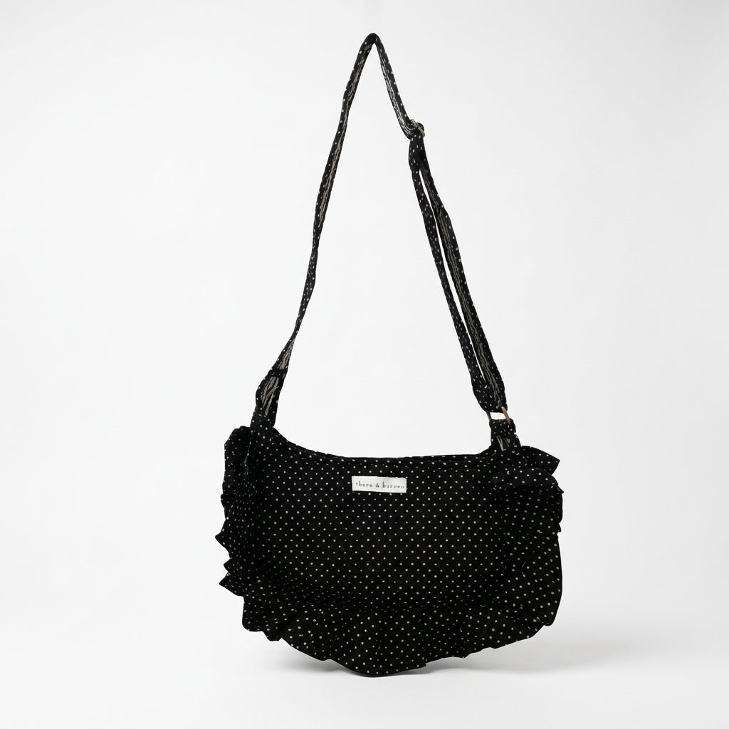 A black fabric shoulder bag with white polka dots hangs from a white metal display stand inside a retail store. 