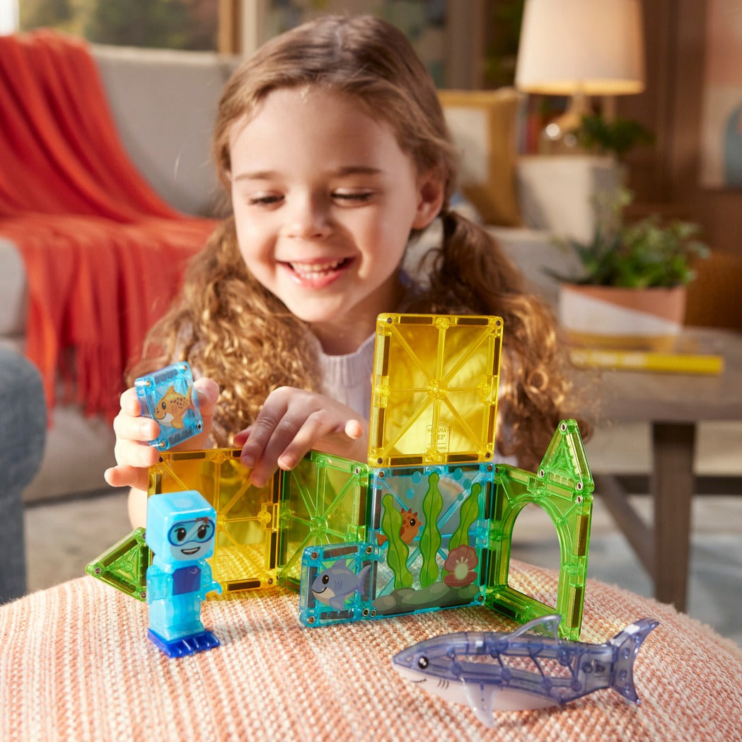 Girl playing with Shark Dive 13-piece magnetic tiles on an ottoman, building an underwater scene with a diver and translucent shark