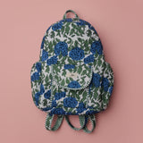 A fabric drawstring backpack featuring a blue rose and green leaf floral pattern on a white background, laid flat against a draped off-white fabric backdrop.